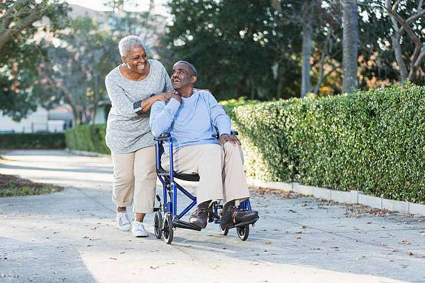 Senior African American couple taking a walk in the park.  The man is in a wheelchair.  His wife is behind, looking down at him and smiling, as she pushes his wheelchair.